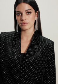 Kvinde med langt mørkt hår iført en sort velvet blazer med små hvide prikker og lange, hængende krystaløreringe, neutral baggrund.