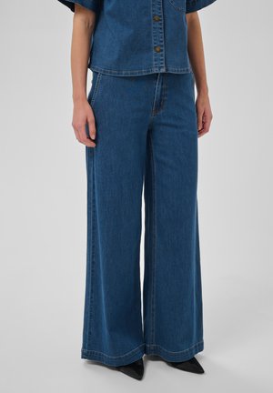 Personne portant une chemise en jean boutonnée et un jean bleu taille haute à jambes larges, mains le long du corps, chaussures noires pointues visibles.