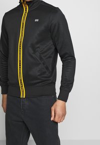 Sort træningsjakke med gul lynlås og branding, featuring standkrave, lange ærmer og sidelommer. Glat stoffinish.