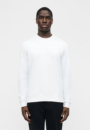 Homme portant un sweat-shirt blanc uni à manches longues et un pantalon noir, debout face à l'avant sur un fond neutre.