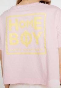 Světle růžové tričko s žlutým textem "HOME BOY" a "LOUD COUTURE" ve čtverci na zadní straně, které nosí osoba s dlouhými blond vlasy.