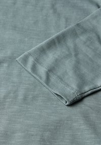 Langärmliges Baumwollkleidungsstück in sanftem Grün, mit einer weichen, strukturierten Oberfläche. Der Ärmelabschluss hat ein sauberes, kontrastierendes Nähdetail.