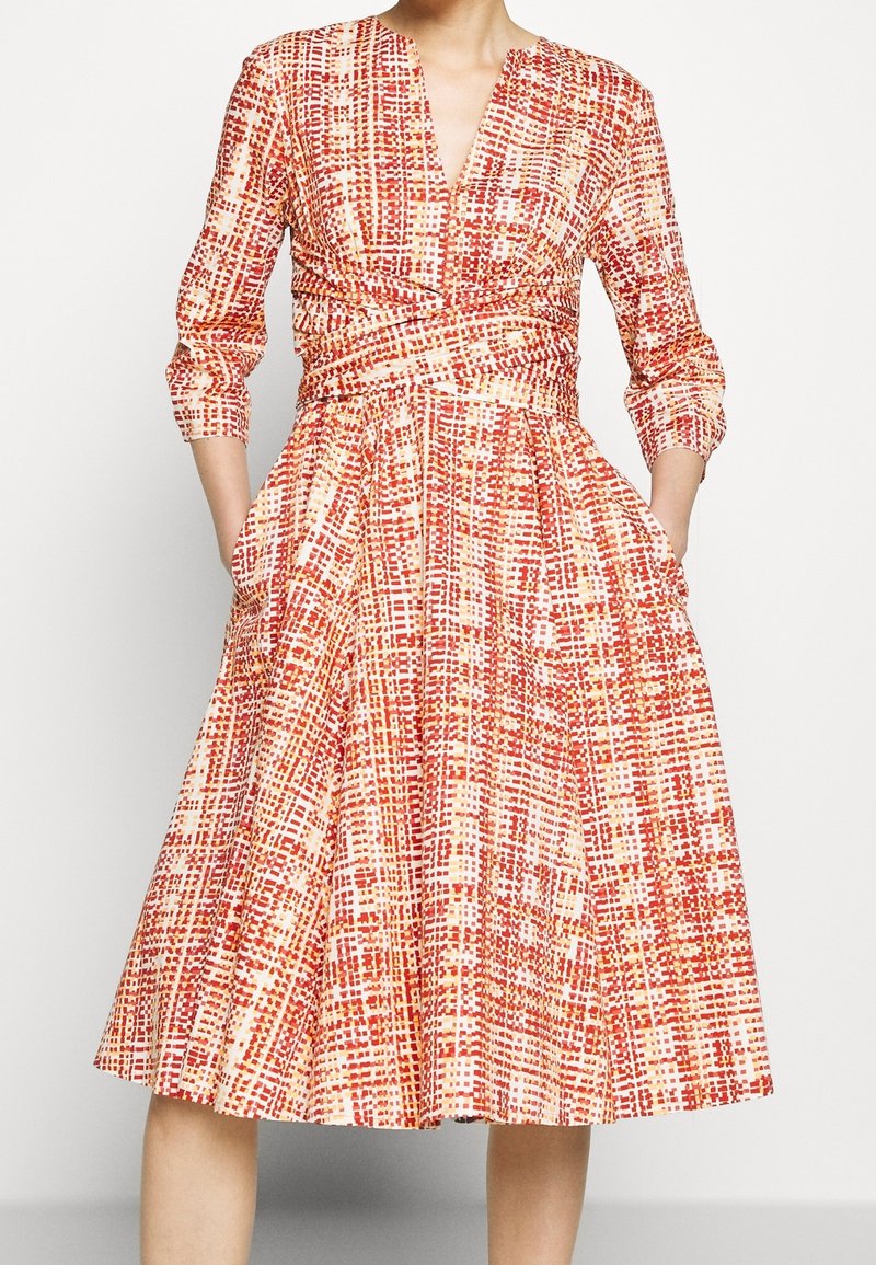 Femme portant une robe mi-longue avec un motif à carreaux rouges, oranges et blancs, des manches trois-quarts, et des poches latérales.