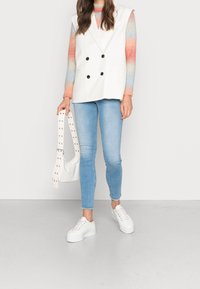 Vit dubbelknäppt väst, ljusblå tajta jeans, vita klumpiga sneakers och en liten vit väska med nitar på remmen. Texturerad regnbågsfärgad tröja.