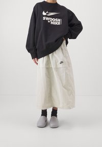 Felpa nera oversize con testo bianco "Swoosh by Nike", abbinata a una gonna beige chiaro con un piccolo logo Nike. Sono indossate delle zoccoli grigi.