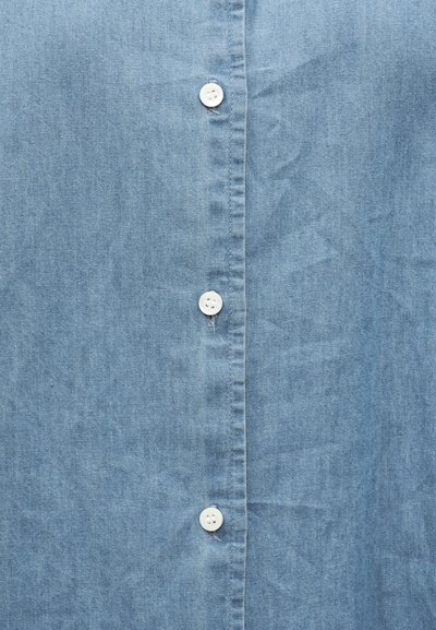 Κοντινό πλάνο ανοιχτού μπλε υφάσματος με τρία λευκά κουμπιά στη σειρά στο μπροστινό μέρος ενός πουκαμίσου ή μπλούζας.