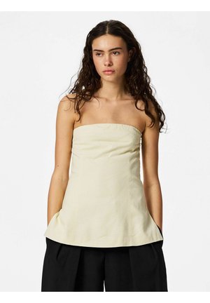 Femme aux longs cheveux bouclés portant un haut beige sans bretelles et un pantalon noir à jambes larges, debout devant un fond blanc uni.