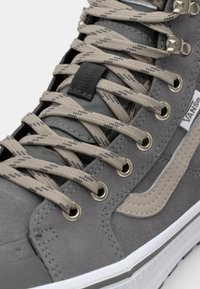 Harmaa korkeavartinen tennari, joka on valmistettu teksturoidusta mokasta ja kankaasta. Tasaiset beige nauhat, metalliset lenkit ja valkoinen kumipohja. Sivussa on logo.