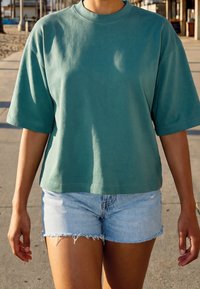 Personne portant une chemise ample verte à manches courtes et un short en jean bleu clair marchant en plein air par une journée ensoleillée.