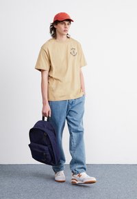 Beige bomulls-t-shirt med grafisk tryck, blå denimjeans, orange keps och marinblå ryggsäck. Vita och orange sneakers med geometrisk design.