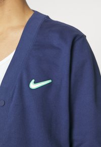 Personne portant un cardigan bleu foncé avec un logo Nike en broderie vert et blanc sur la poitrine.