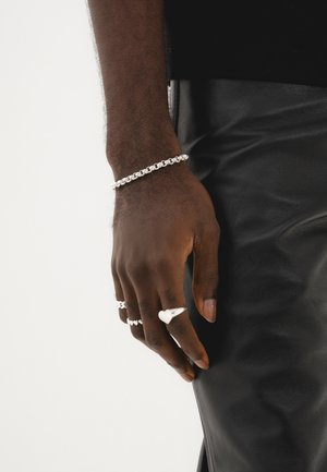 Silberne Kettenarmbanduhr und drei Ringe an einer Hand, wobei einer der Ringe herzförmig ist. Die Hand liegt auf einer schwarzen Lederoberfläche.