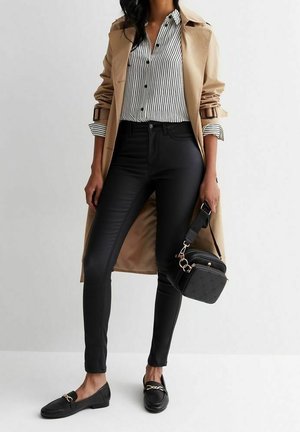 Kvinde iført beige trenchcoat, sorte smalle bukser, stribet skjorte med knapper, sorte loafers og bærende en sort quiltet crossbodytaske.