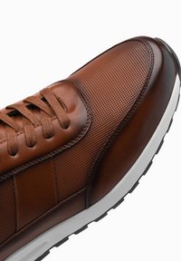 Bruine leren sneaker met een geperforeerde textuur, vetersluiting, contrasterende witte zool en zwarte rubberen loopzool.