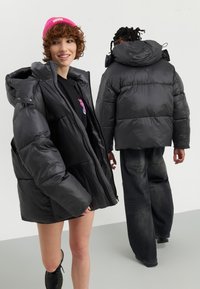 Svart puffjakke med blank finish, stor hette og frontglidelås. Bært over en svart skjorte, sammen med svarte jeans og støvler.
