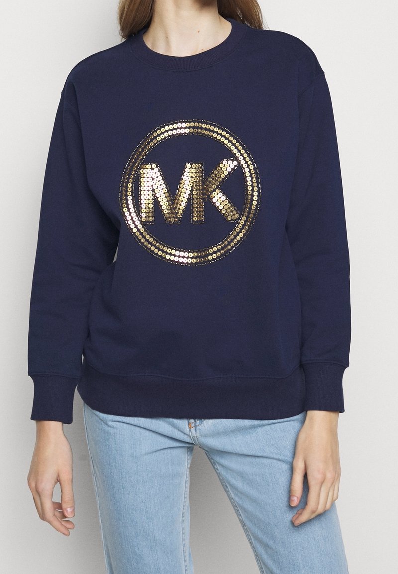 Personne portant un sweat-shirt bleu marine avec un grand cercle de sequins dorés « MK » sur la poitrine, associé à un jean bleu clair.