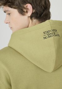 Sweat à capuche couleur olive avec une texture douce, arborant une grande capuche au dos et un texte brodé noir : "RESTEZ OUVERT, IL N'Y A PAS DE MAUVAIS JOURS."