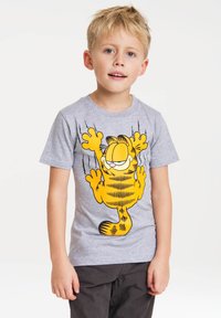 Šedé bavlněné tričko se vzorem kreslené kočky žluté barvy s černými pruhy, stojící s raise paw na čistém pozadí.