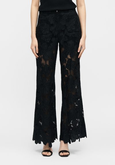 Γυναίκα που φοράει μαύρο δαντελένιο παντελόνι με φαρδιά μπατζάκια με σχέδιο λουλουδιών και μαύρα σανδάλια με ανοιχτές μύτες, στέκεται μπροστά σε απλό φωτεινό φόντο.