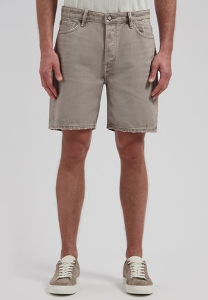 Mann trägt beige Jeansshorts und taupefarbene Sneakers mit weißen Schnürsenkeln, steht vor schlichtem hellgrauem Hintergrund.