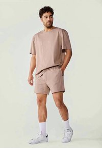 T-shirt de algodão de manga curta, cor castanho claro, combinado com calções correspondentes. Apresenta um corte descontraído, bolsos laterais e meias desportivas brancas com sapatilhas.