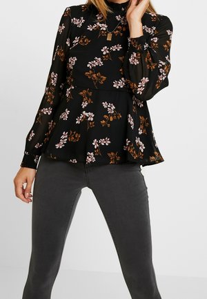Vrouw draagt een zwarte bloemenblouse met lange mouwen, geplooide manchetten en donkergrijze aansluitende jeans, poseert met één hand bij haar gezicht.