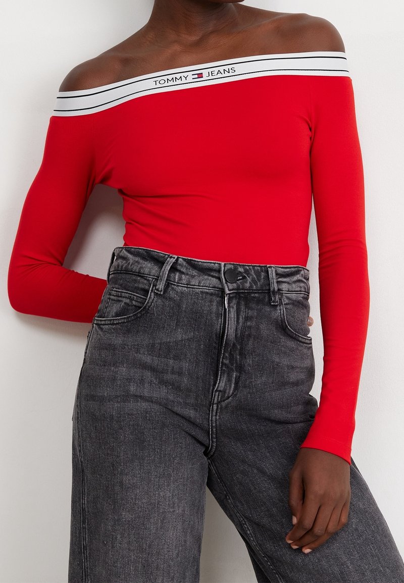 Henkilö, joka on pukeutunut kirkkaan punaiseen olkaimeton pitkähihaiseen puseroon, jossa on valkoinen Tommy Jeans -logo, yhdistettynä korkeavyötäröisiin haalistuneisiin mustiin farkkuihin.