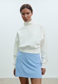 Biely skrátený sveter s vysokým golierom; kombinovaný so svetlomodrou minisukňou. Hladká látka, voľný strih a minimalistický dizajn.
