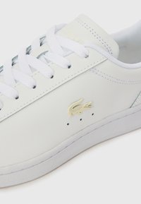 Hvite sneakers laget av glatt lær med hvite lisser, en gulgrønn logo og subtile perforeringer nær logoen, med gummisåle.