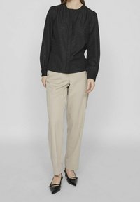 Donna che indossa una blusa nera a maniche lunghe con pattern floreali sottili, pantaloni beige a gamba dritta e scarpe con tacco a punta nere.