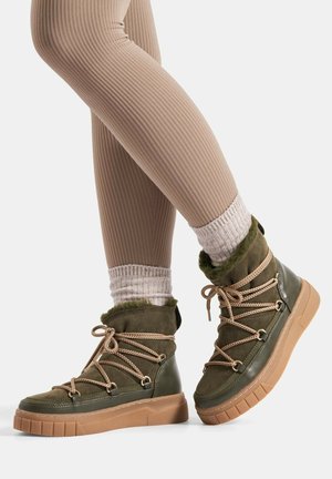 Nogi w beżowych prążkowanych legginsach i jasnoszarych skarpetkach, noszące oliwkowozielone, futrzane, sznurowane buty na grubej brązowej podeszwie.