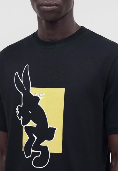 Personne portant un t-shirt noir avec une grande silhouette noire de lapin délimitée en blanc sur un rectangle jaune au niveau de la poitrine.