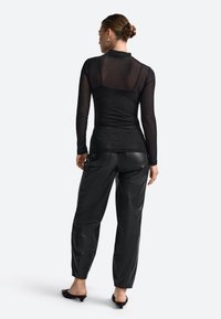 Top negru cu mâneci lungi, cu panouri transparente și textură strălucitoare, asortat cu pantaloni negri cu talie înaltă și lucioși și tocuri negre.