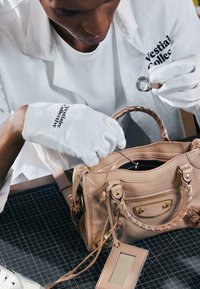Personne portant des gants blancs inspecte un sac à main en cuir beige avec une loupe et un outil sur un tapis de découpe.