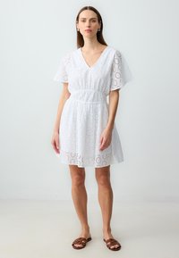 Weißes Baumwollkleid mit Lochstickerei, V-Ausschnitt, kurzen Flugärmeln und elastischem Bund, das eine fließende A-Linie Silhouette bietet.