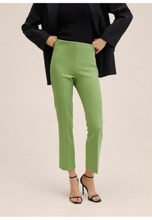 Mujer vistiendo pantalones verdes ajustados, blusa negra acanalada, blazer negro y sandalias negras de tacón alto, de pie con las manos en la cadera.