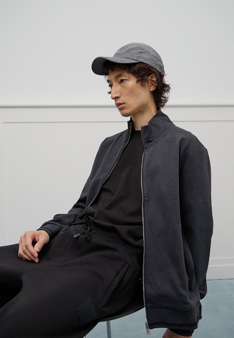 Donkergrijze zip-up jas over een zwart shirt en broek, met een lichtgrijze pet. Heeft een relaxte pasvorm en een zachte textuur. Minimalistisch ontwerp.