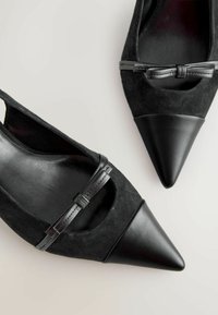 Pantofi cu toc ascuțit negri, realizați din suede și piele, cu un design cu decupaj și un accent de fundiță mică cu un finisaj lucios pe vârf.