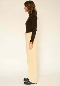 Haut à manches longues marron à côtes associé à un pantalon plissé beige clair, avec une coupe droite et des chaussures à accent noir, vu de côté.