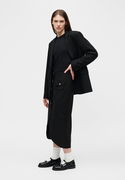 Γυναίκα που ποζάρει φορώντας μαύρο σακάκι, μαύρο τοπ, μαύρη μίντι φούστα με κουμπιά και τσέπες, λευκές κάλτσες και μαύρα δερμάτινα παπούτσια, σε απλό φόντο.