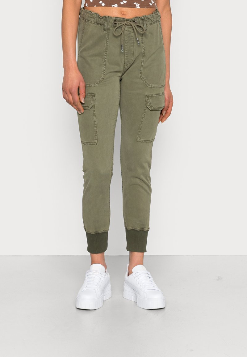Femme portant un pantalon cargo jogger vert olive avec taille à cordon et poignets côtelés, associé à des baskets plateformes blanches.
