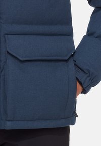 Donkerblauwe pufferjack met een klepzak, geribbelde manchetten en een gestructureerde stof. Het ontwerp is gevoerd voor warmte en comfort.