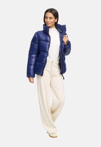 Niebieska kurtka puchowa z wysokim kołnierzem, błyszczącą fakturą i pionowym pikowaniem. W komplecie z kremowymi spodniami o szerokich nogawkach i jasnym obuwiem.