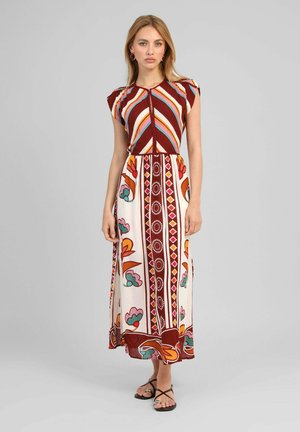 Jeune femme portant une robe longue sans manches à motifs géométriques et floraux, debout avec une expression neutre devant un fond uni.