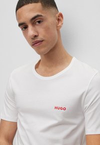 T-shirt en coton blanc avec un col rond, présentant un logo "HUGO" rouge brodé sur la poitrine gauche. Texture douce et manches courtes.