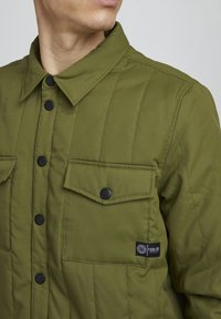 Giacca da camicia imbottita verde oliva con bottoni a pressione neri, due tasche sul petto e un'etichetta; presenta una trama trapuntata e un colletto.