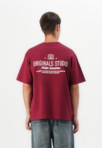 Bordeauxrød bomulds T-shirt med rund halsudskæring, der har hvid tekst og et grafisk logo på bagsiden. Sammen med blå denim jeans.