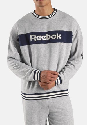 Mann trägt grauen Reebok-Sweatshirt mit marineblauem Streifen und weißem Logo, passende graue Hose, Hände vor der Taille verschränkt.