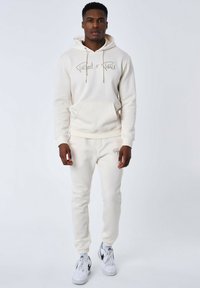 Felpa con cappuccio color crema con tasca anteriore e logo in corsivo, abbinata a pantaloni sportivi tapered coordinati, entrambi in un materiale morbido.