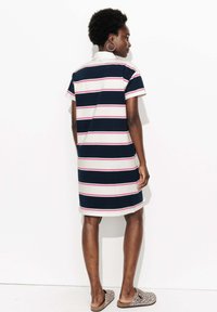 Femme se tenant face à un mur blanc, portant une robe rayée bleu marine et blanche avec des accents roses et des chaussures à enfiler à imprimé léopard.